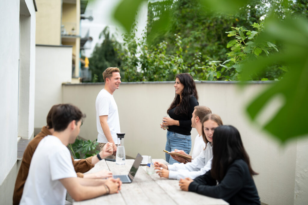 Eine Gruppe von sechs jungen Erwachsenen, die in einer freundlichen und entspannten Atmosphäre draußen zusammenkommen. Sie befinden sich in einem offenen, halb überdachten Bereich, umgeben von grüner Vegetation und modernen Gebäuden im Hintergrund.