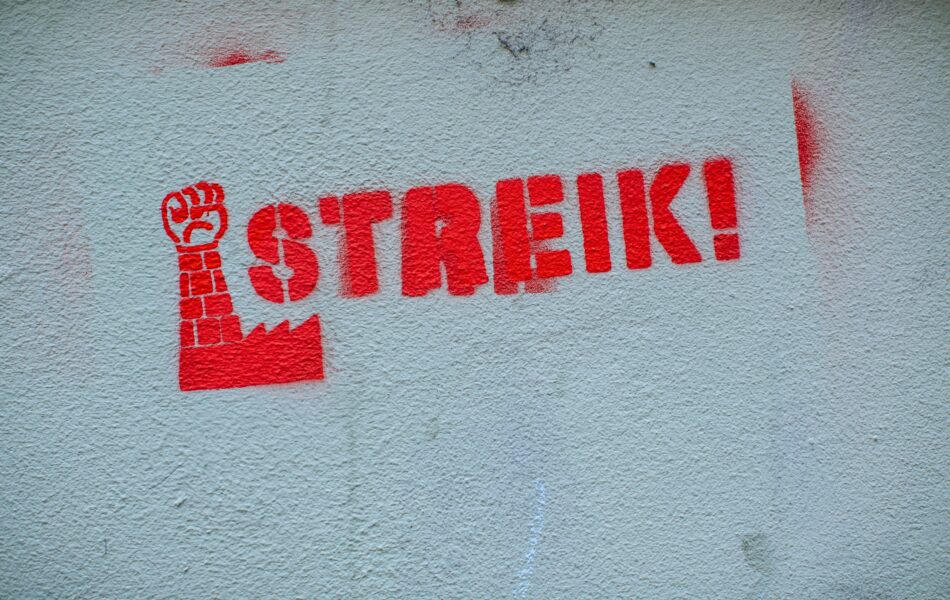 Ein Wandgraffiti mit dem Wort “STREIK!” in roter Farbe. Links neben dem Schriftzug ist ein Symbol zu sehen: eine rote Faust, die nach oben gereckt ist und deren Arm aus Ziegeln besteht. Der Arm ragt aus einem stilisierten Fabrikgebäude empor. Dieses Bild vermittelt eine klare Botschaft des Protests und der Arbeitersolidarität. Der Schriftzug und das Symbol sind beide in einem kräftigen, auffälligen Rot gehalten, was die Dringlichkeit und den Nachdruck des Themas eines Streik unterstreicht.