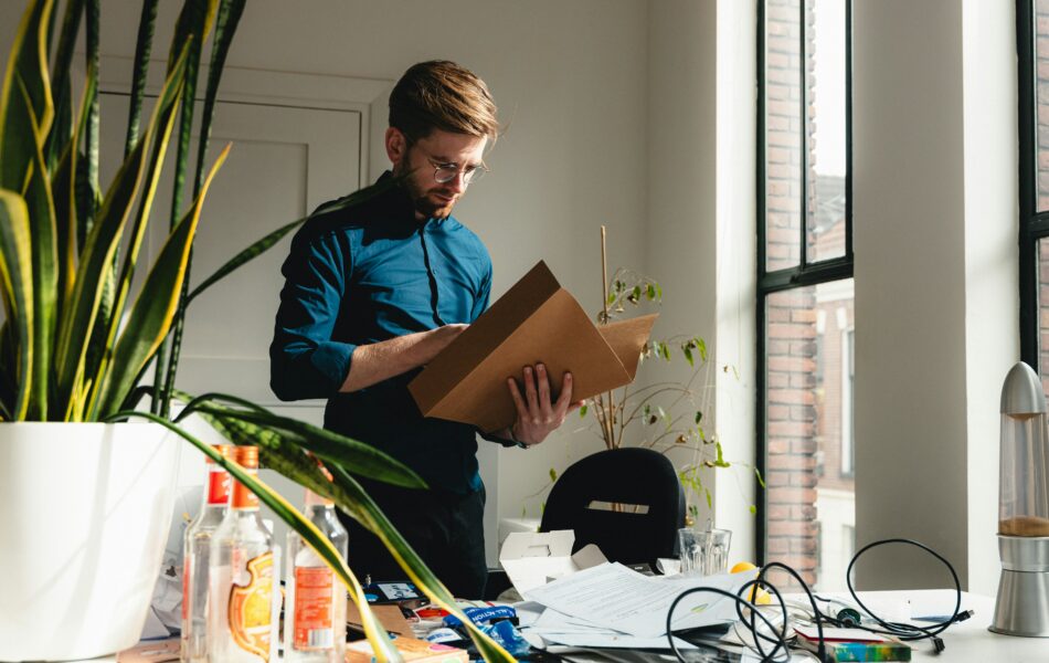 Ein Mann steht konzentriert in einem hellen Büro, eine geöffnete Aktenmappe in den Händen, während sein Blick auf die darin befindlichen Dokumente gerichtet ist. Der Schreibtisch vor ihm ist von Papieren, leeren Flaschen und Kabeln bedeckt, was eine hektische oder unorganisierte Arbeitsumgebung widerspiegelt. Im Zusammenhang mit Personalstammdaten symbolisiert das Bild die Notwendigkeit, Struktur und Ordnung bei der Erfassung und Verwaltung von Mitarbeiterdaten zu schaffen, um Unübersichtlichkeit und Fehler zu vermeiden.