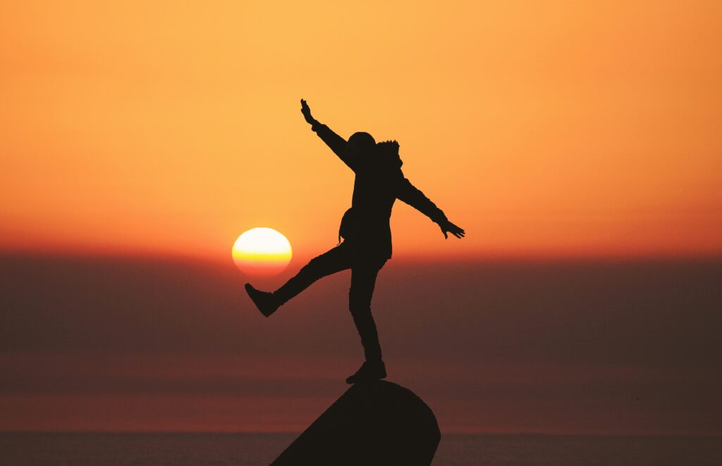 Eine Person balanciert auf einem Vorsprung, die Silhouette zeichnet sich vor einem leuchtenden Sonnenuntergang ab. Die ausgestreckten Arme und das Gleichgewicht auf einem Bein symbolisieren die Balance zwischen Herausforderung und Stabilität. Studienbegleitetes Arbeiten verweist hier auf die Notwendigkeit, Studium und berufliche Tätigkeiten geschickt miteinander zu vereinen, um eine gesunde Balance zwischen Lernen, Arbeiten und persönlicher Entwicklung zu erreichen.