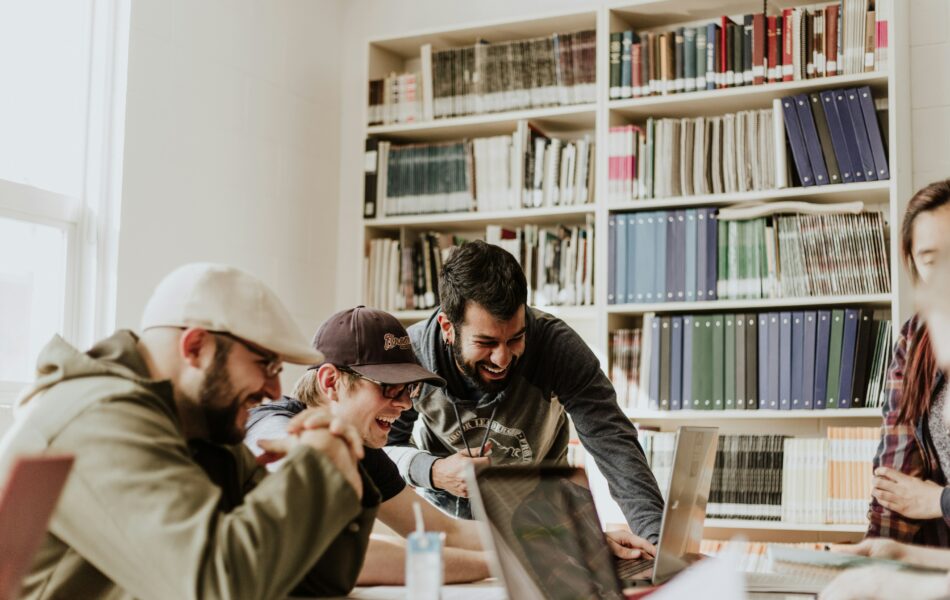 Eine Gruppe junger Menschen sitzt in einem hellen Raum an einem Tisch, arbeitet gemeinsam an Laptops und zeigt Freude und Begeisterung. Bücherregale im Hintergrund deuten auf einen Lern- oder Studienkontext hin. Studienbegleitetes Arbeiten verdeutlicht hier die Möglichkeit, Studium und praktische Teamarbeit zu kombinieren, um Wissen effektiv anzuwenden und wertvolle berufliche Erfahrungen zu sammeln.