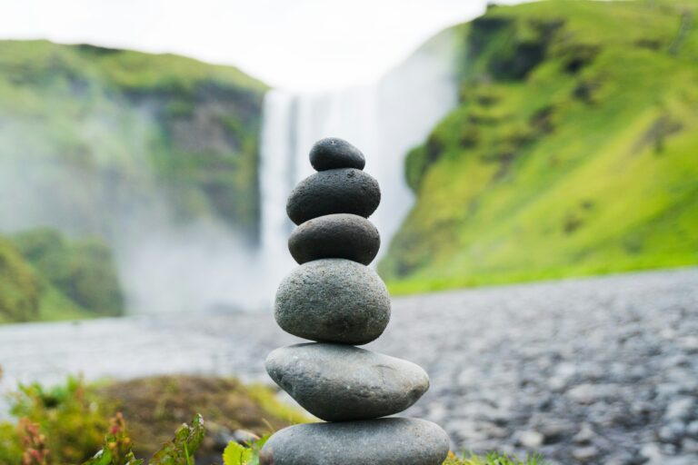 Ein Stapel sorgfältig ausbalancierter Steine steht vor einem malerischen Wasserfall, umgeben von grünen Hügeln. Das Bild symbolisiert Gleichgewicht und Präzision, die bei komplexen Prozessen erforderlich sind. Lohnkorrektur verweist hier auf die Notwendigkeit, Ungleichgewichte in der Lohnabrechnung auszugleichen und sicherzustellen, dass Fehler präzise behoben werden, um die finanzielle Genauigkeit und Fairness zu wahren.