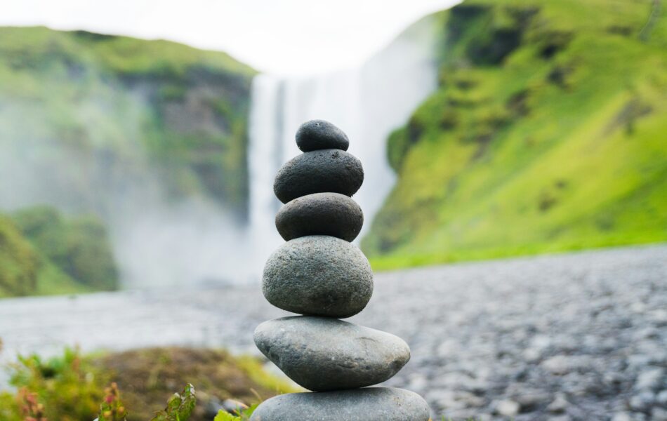 Ein Stapel sorgfältig ausbalancierter Steine steht vor einem malerischen Wasserfall, umgeben von grünen Hügeln. Das Bild symbolisiert Gleichgewicht und Präzision, die bei komplexen Prozessen erforderlich sind. Lohnkorrektur verweist hier auf die Notwendigkeit, Ungleichgewichte in der Lohnabrechnung auszugleichen und sicherzustellen, dass Fehler präzise behoben werden, um die finanzielle Genauigkeit und Fairness zu wahren.
