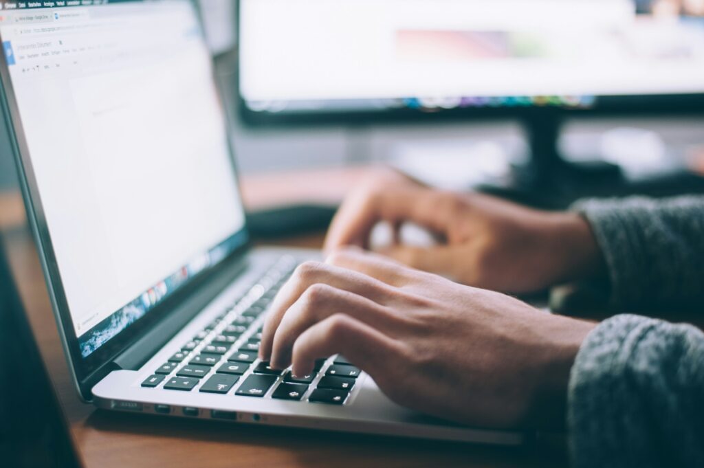 Eine Person tippt konzentriert auf einem Laptop, während im Hintergrund ein zweiter Bildschirm zu sehen ist. Die Szene vermittelt eine professionelle Arbeitsumgebung, in der Daten sorgfältig bearbeitet und überprüft werden. SFN-Zuschläge (Sonn-, Feiertags- und Nachtzuschläge) verweisen hier auf die Rolle der Lohnbuchhaltung, die diese Zuschläge korrekt berechnen und erfassen muss, um die gerechte Vergütung für Arbeitsleistungen zu besonderen Zeiten sicherzustellen.